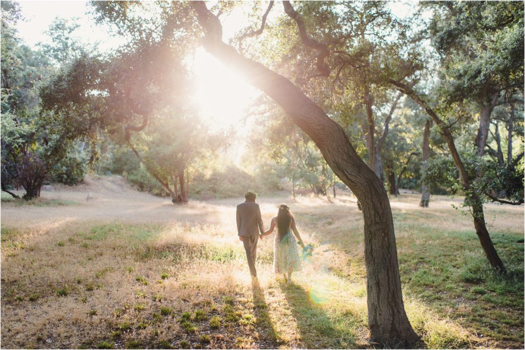 Erica and Kevin Hold hands as they walk through oak trees during Golden hour at their whimsical Garden Wedding