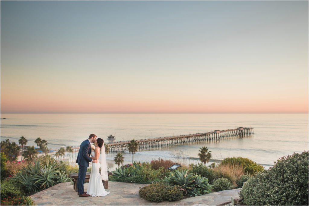 A bride and groom kiss on a patio at Casa Romantica overlooking the San Clemente Pier on the Pacific Ocean at their Southern California Cliffside Wedding