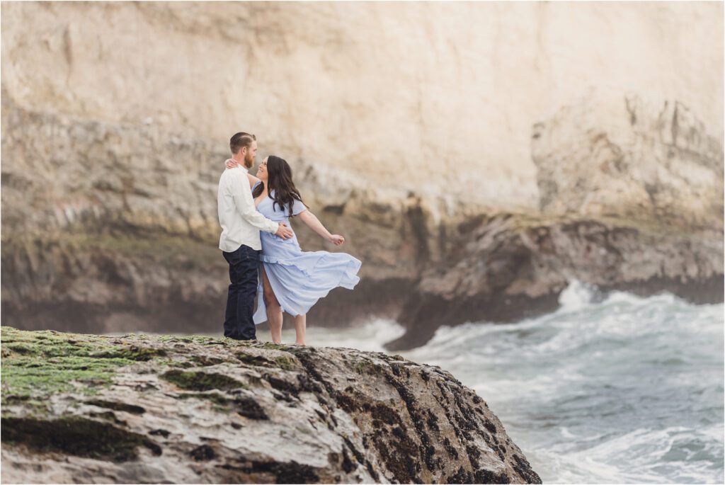 A couple poses on a rock during their shark fin cove engagement session with Sun & Sparrow