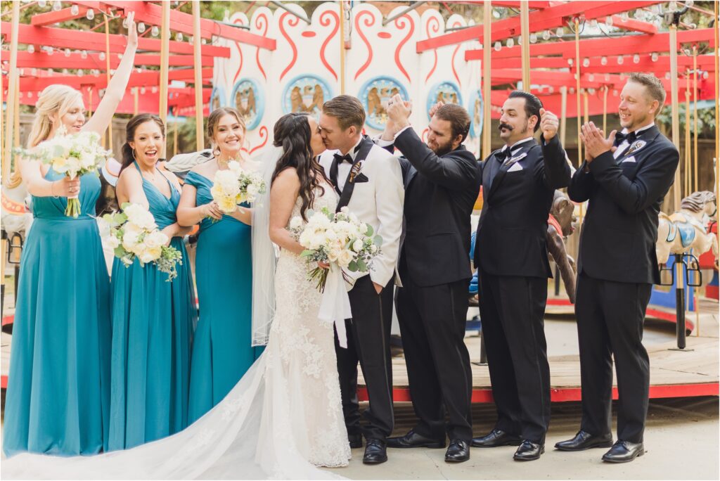 the wedding party cheers while a bride and groom kiss in front of the merry go round during a calamigos ranch birchwood room wedding