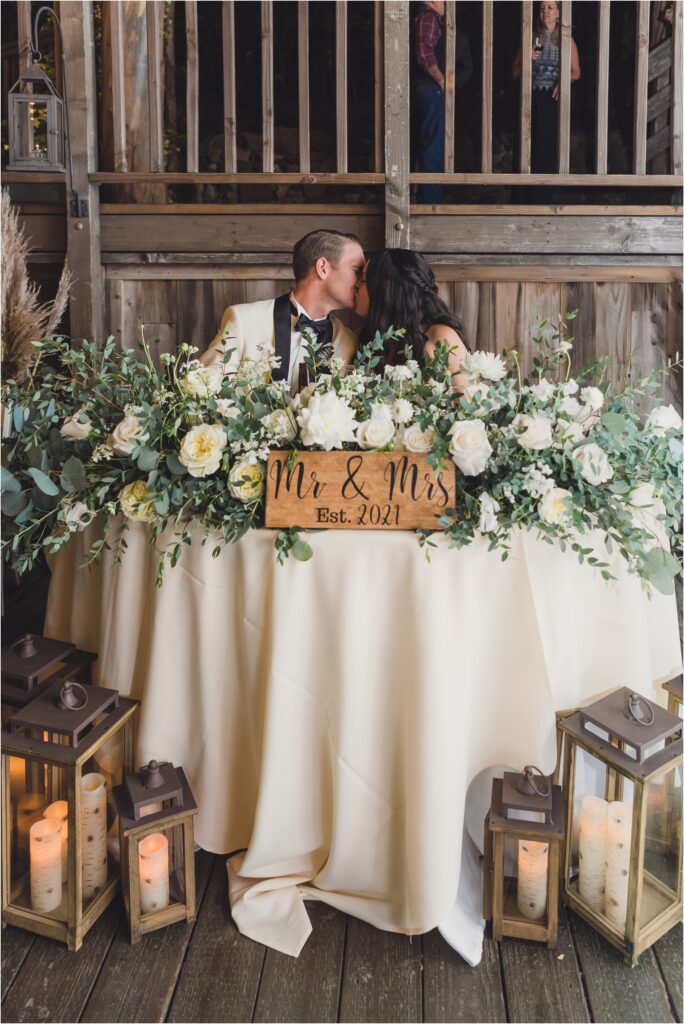 Sitting at their sweetheart table covered in white flowers, a bride and groom kiss during their Calamigos Ranch Birchwood room wedding