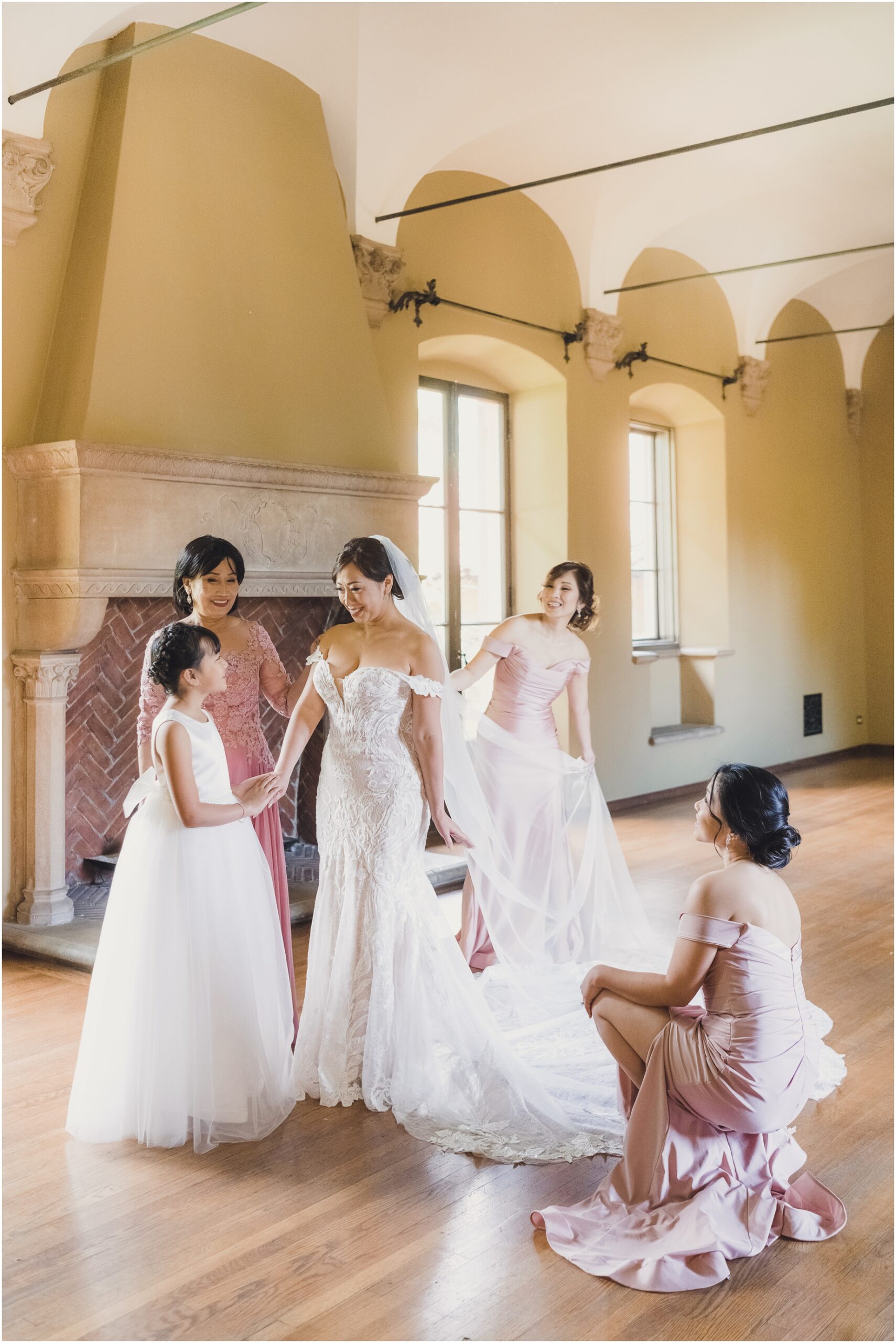 A bride and her bridesmaids getting ready for a wedding at Villa del sol d'Oro. The bride is looking at her daughter with love, while her bridesmaids look on. The room has a large fireplace with brick and stone, while the walls have arches and the floors are hardwood