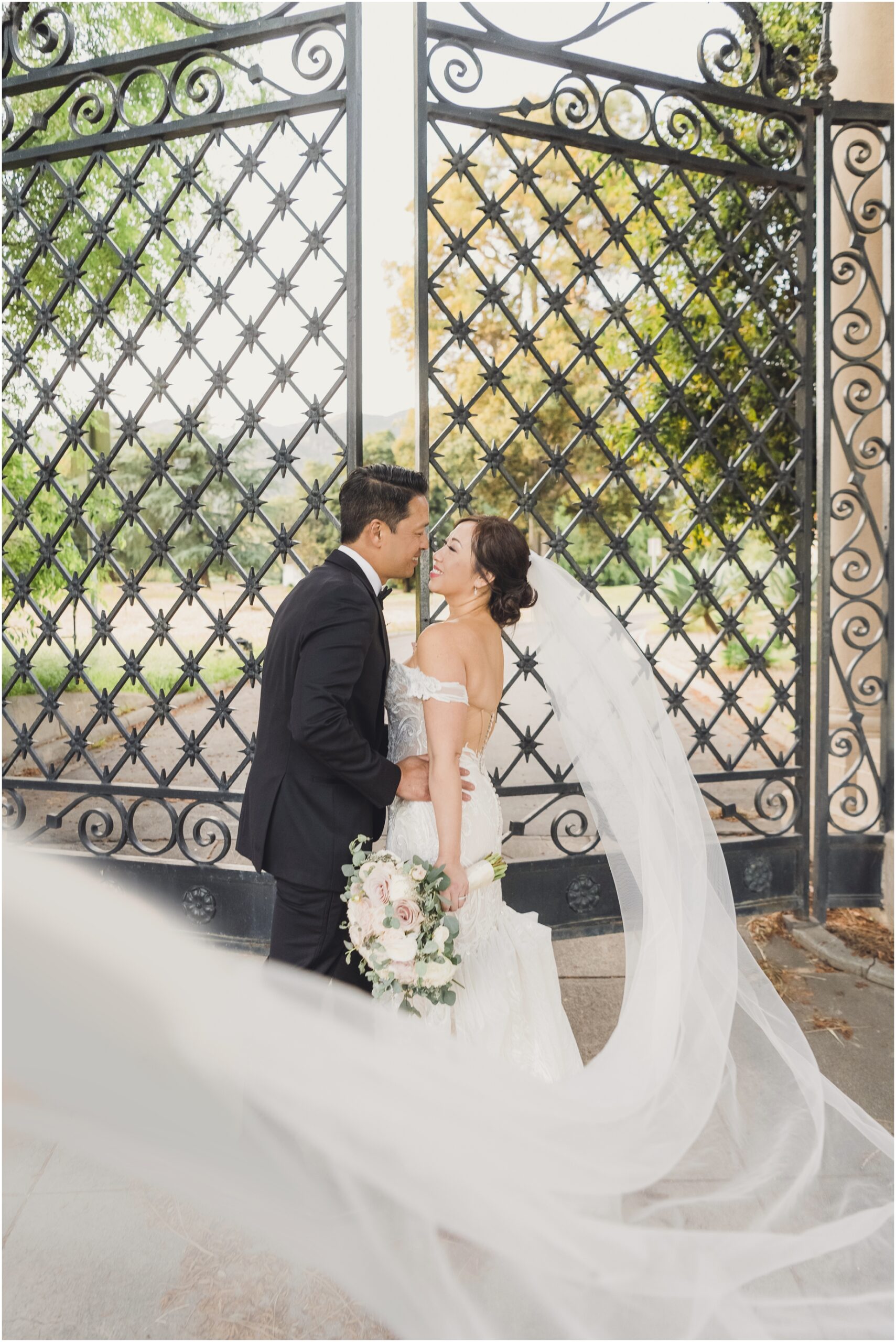 A bride and groom pose in front of an opening gate at Villa del sol d'Oro. the bride is holding a pink and white bouquet as her veil crosses the bottom of the photograph.