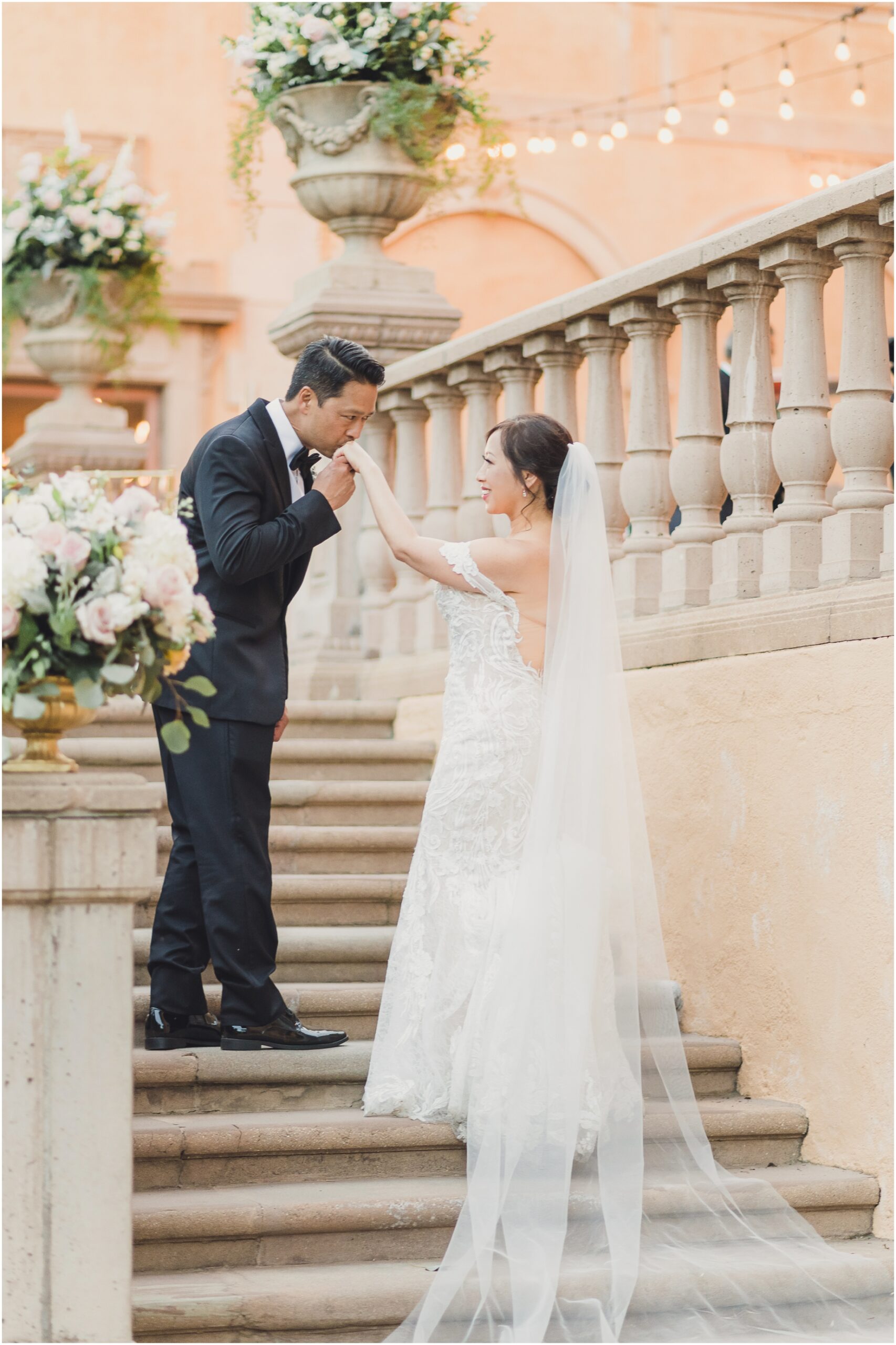 A groom kisses the hand of his bride on the steps below their wedding reception at Villa del sol d'Oro. The bride has a gorgeous veil that trails all the way down the stairs. photo taken by sun and sparrow photography