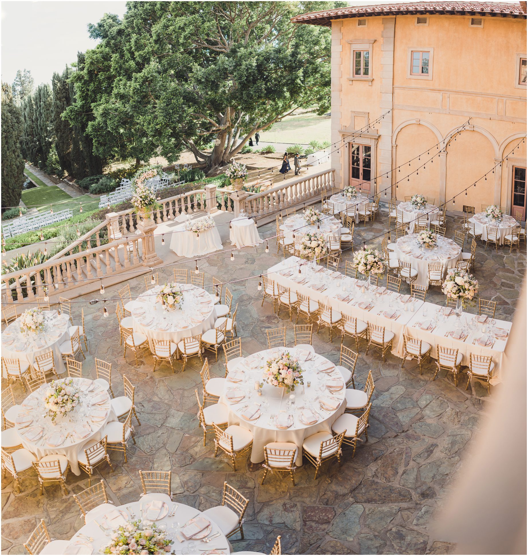 A wedding reception setup on a balcony at a gorgeous Villa del sol d'orzo wedding. The wedding features gold chairs, pink and white flowers, hanging lights, and white tablecloths