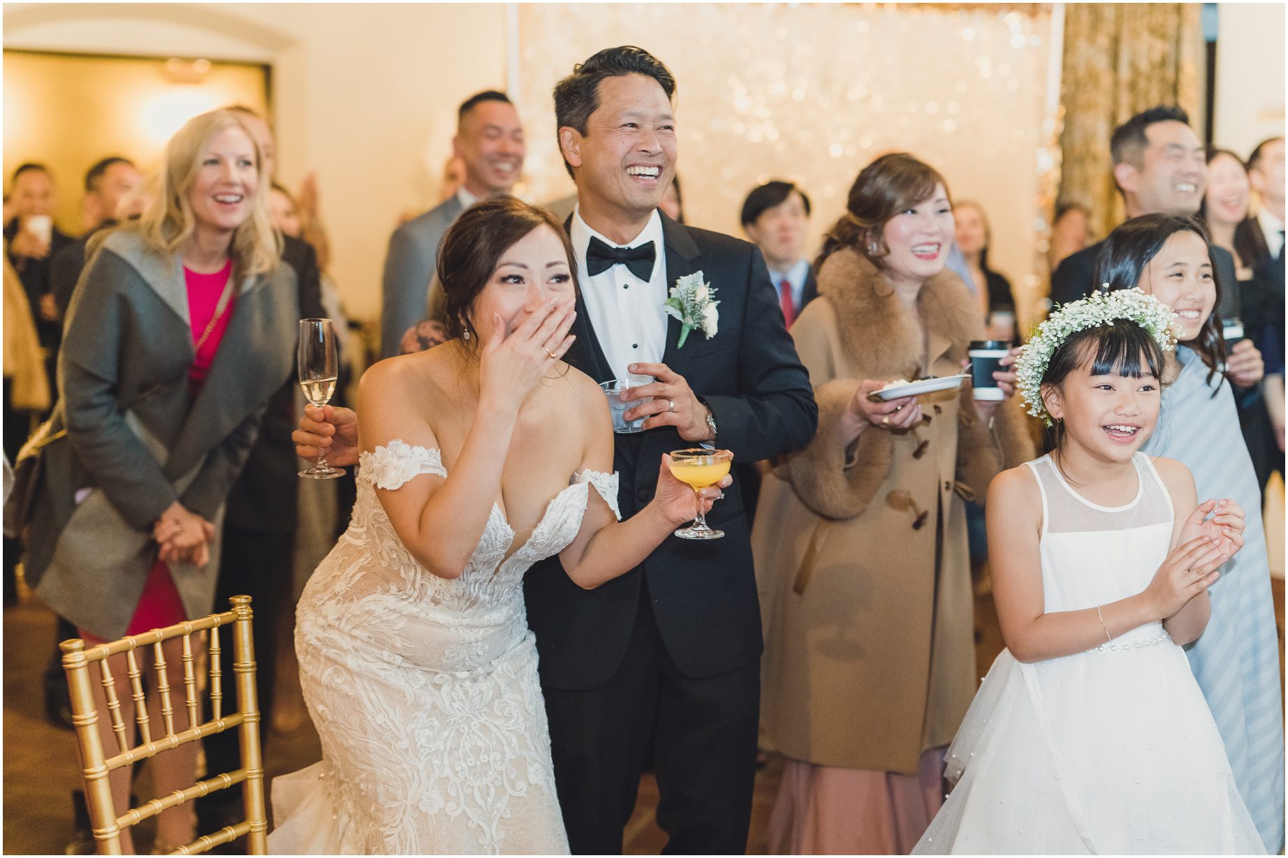 A bride leans and covers her mouth in amusement while the groom smiles next to her and other people observe the same thing