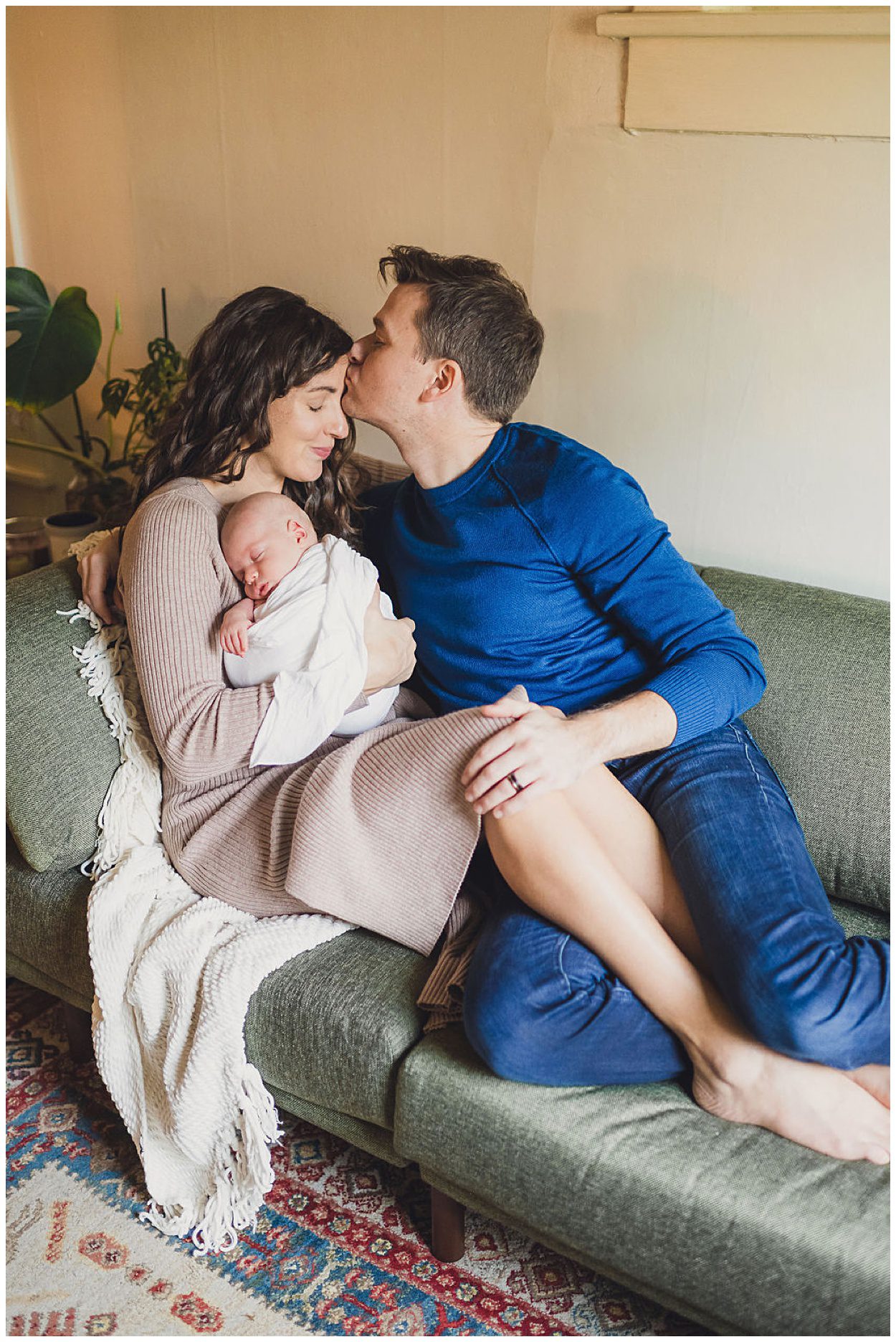 A mother and father cuddle on the couch while holding their newborn child in Portland Oregon. The father is kissing the mother on the forehead and the baby is sleeping in the mother's arms