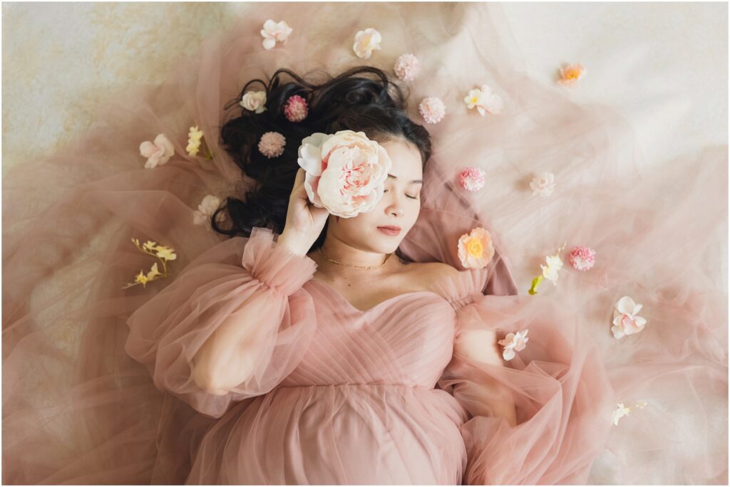 At her studio Maternity session in Portland, an expecting mother poses with pink, white, and peach flowers, while wearing a flowy dress and laying on a bed of fabric. Sun and Sparrow is a Portland Maternity and Newborn Photographer