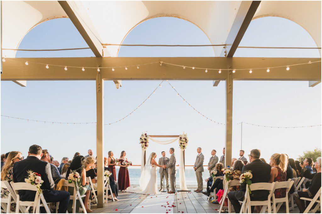 A wedding on the beach at Malibu west beach club, featuring a white arch and wooden chairs
