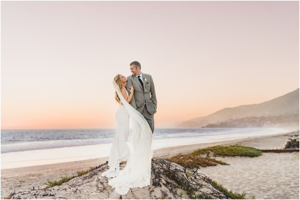 A bride in an Enzoani dress and a groom pose on the beach during their sunset beach wedding Malibu West Beach Club