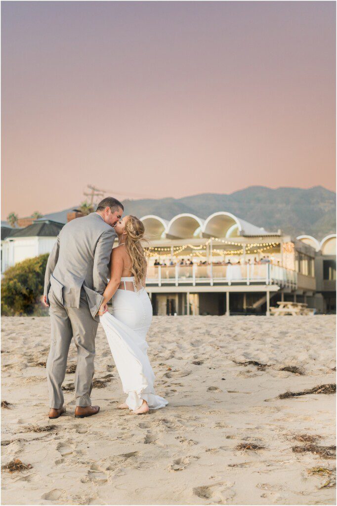 A bride and groom kiss at sunset on the beach in Malibu, in front of Malibu west beach club