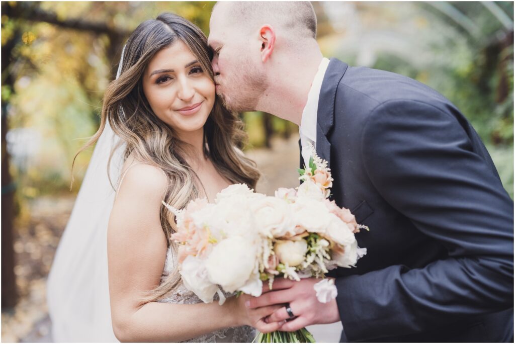 A bride looks at the camera and holds her bouquet as the groom kisses her cheek gently