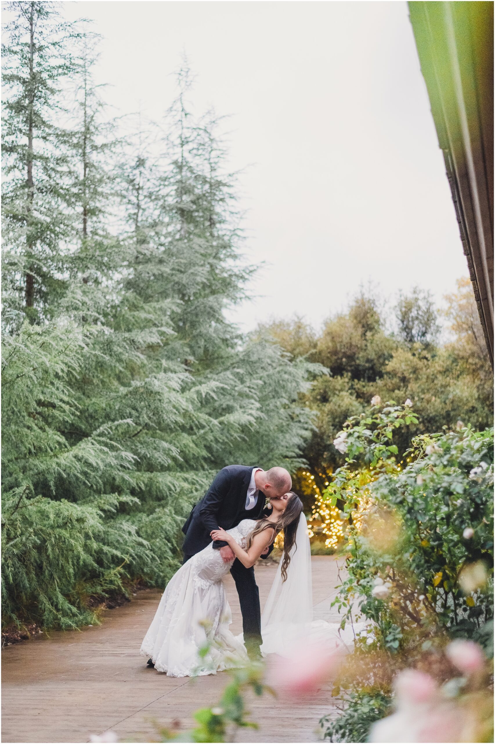 A groom dips his bride during couples portraits with Sun & Sparrow at their Serendipity Gardens wedding. the picture features evergreens and a garden