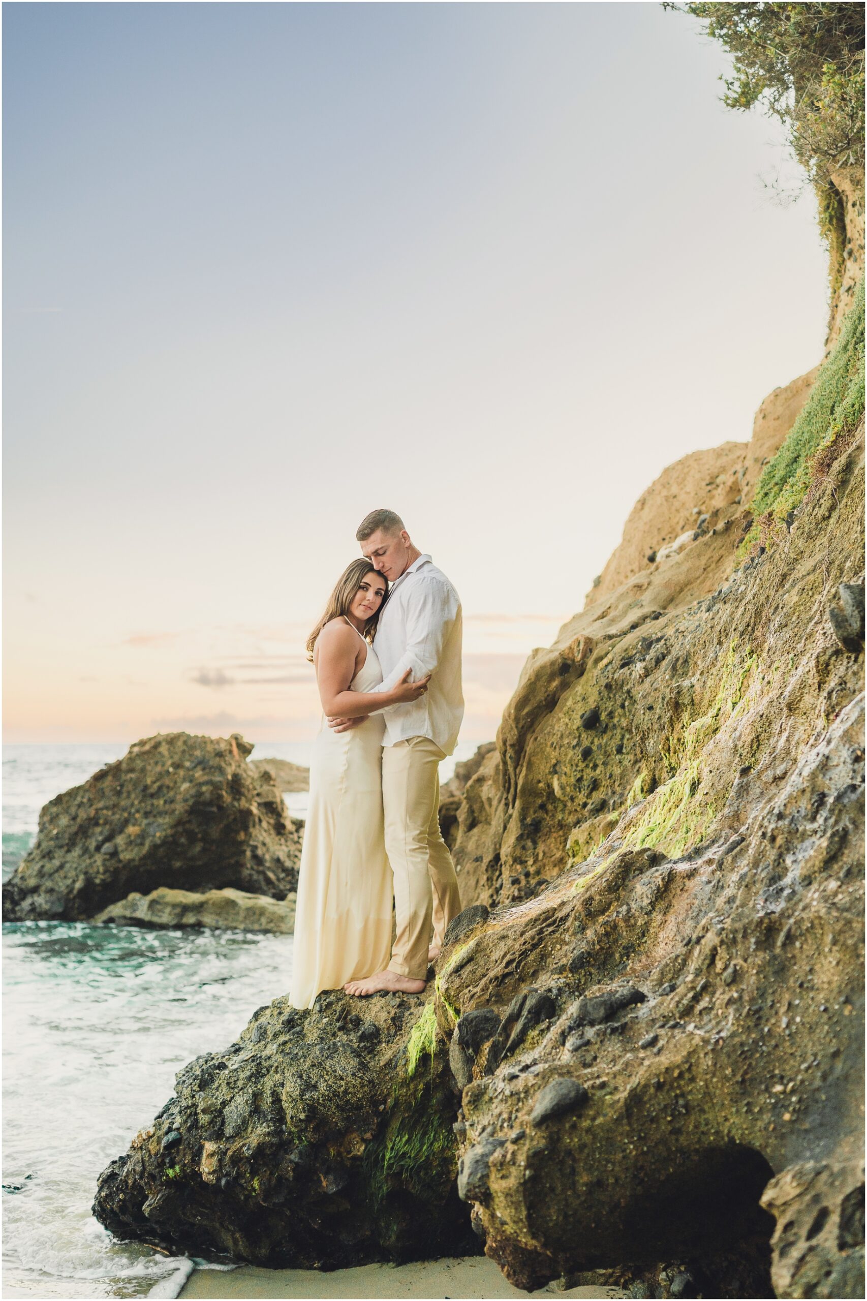 Couples portrait at Woods Cove in Laguna Beach featuring a woman leaning into a man while standing on a rocky outcropping over the ocean