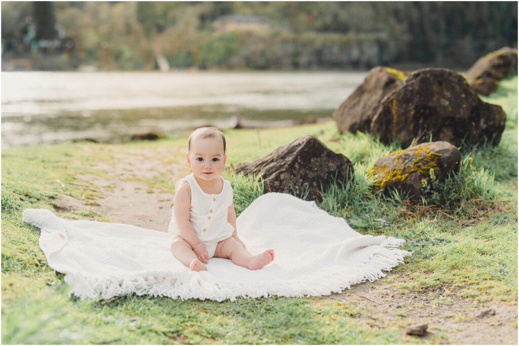 A one year old boy sitting on a white blanket near the Willamette River in Oregon during a Lake Oswego Family Photo Session