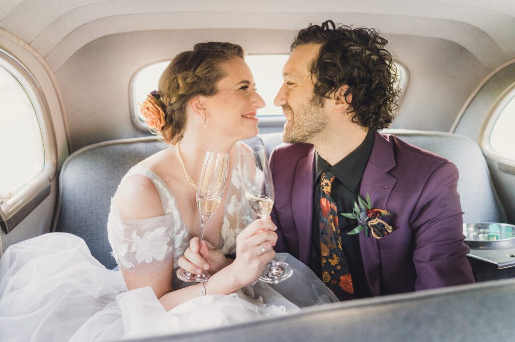 A bride and groom sit in the backseat of a classic car, sipping champagne while looking into each others eyes during their Solar Eclipse Wedding