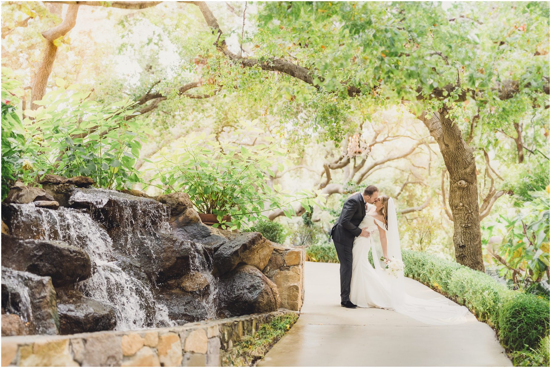 A bride and groom on a path at the oak room, kissing next to a waterfall